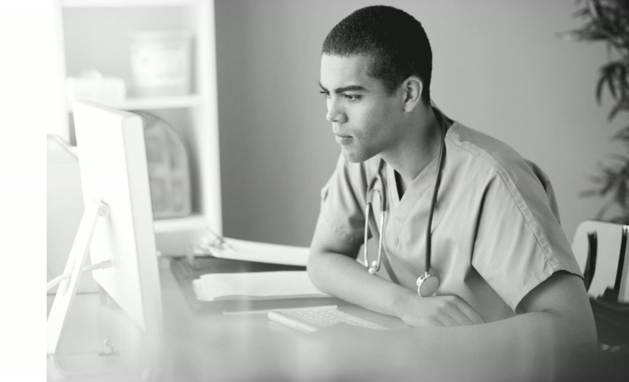A care worker in scrubs with a stethoscope, sitting at a desk and reading a desktop screen, paperwork beside the keyboard. Black and white photograph.