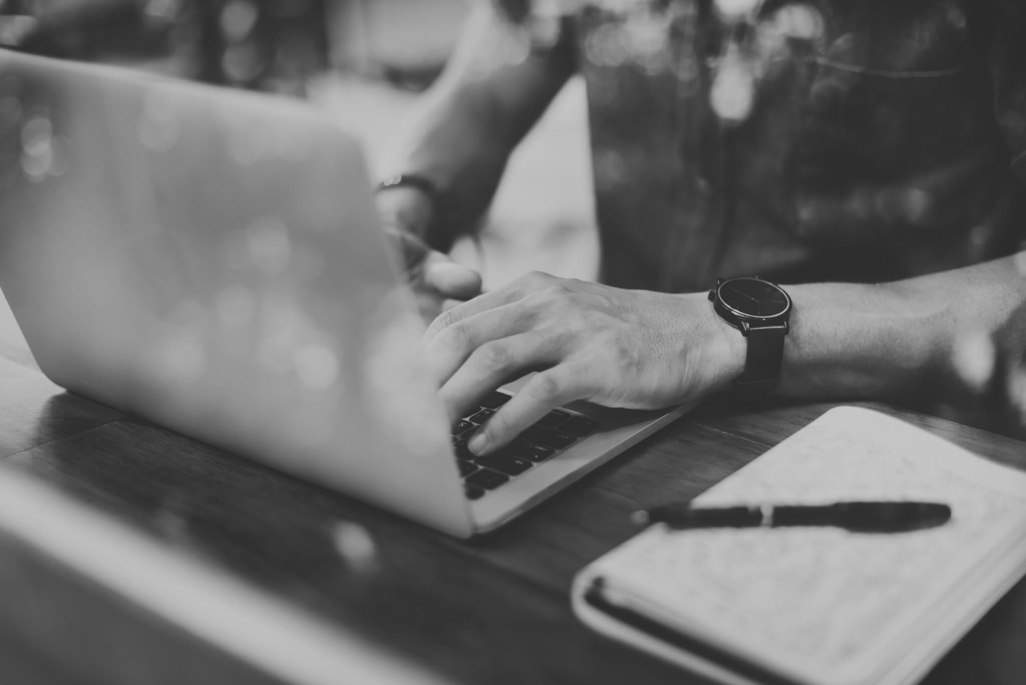 Close-up of hands typing on a laptop at a wooden table, an open notebook and a pen beside it. Black and white photograph.