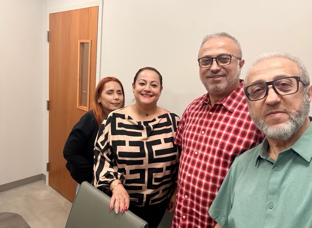 Four people posing together indoors in front of a light wooden door.