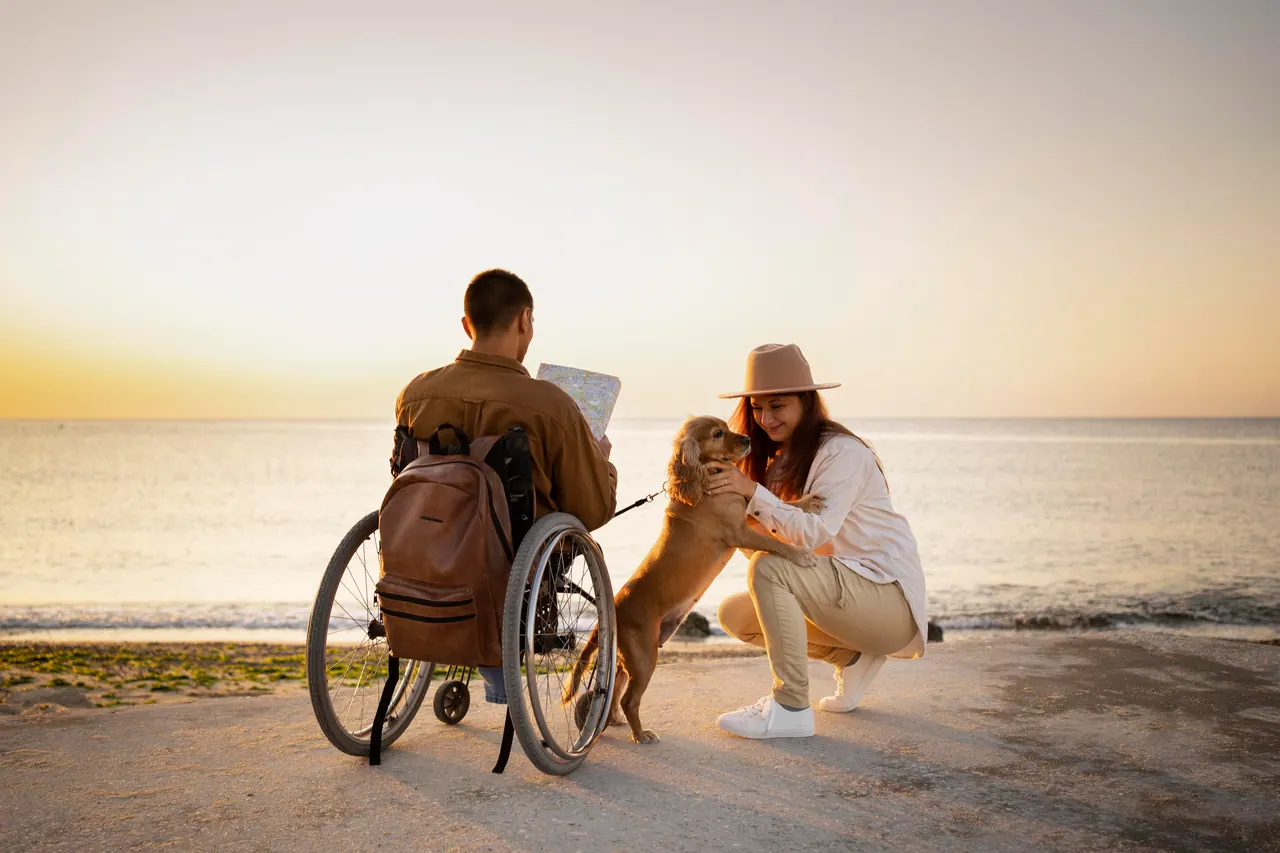 A wheelchair user and a companion stop with their dog on a path by the sea at sunset.